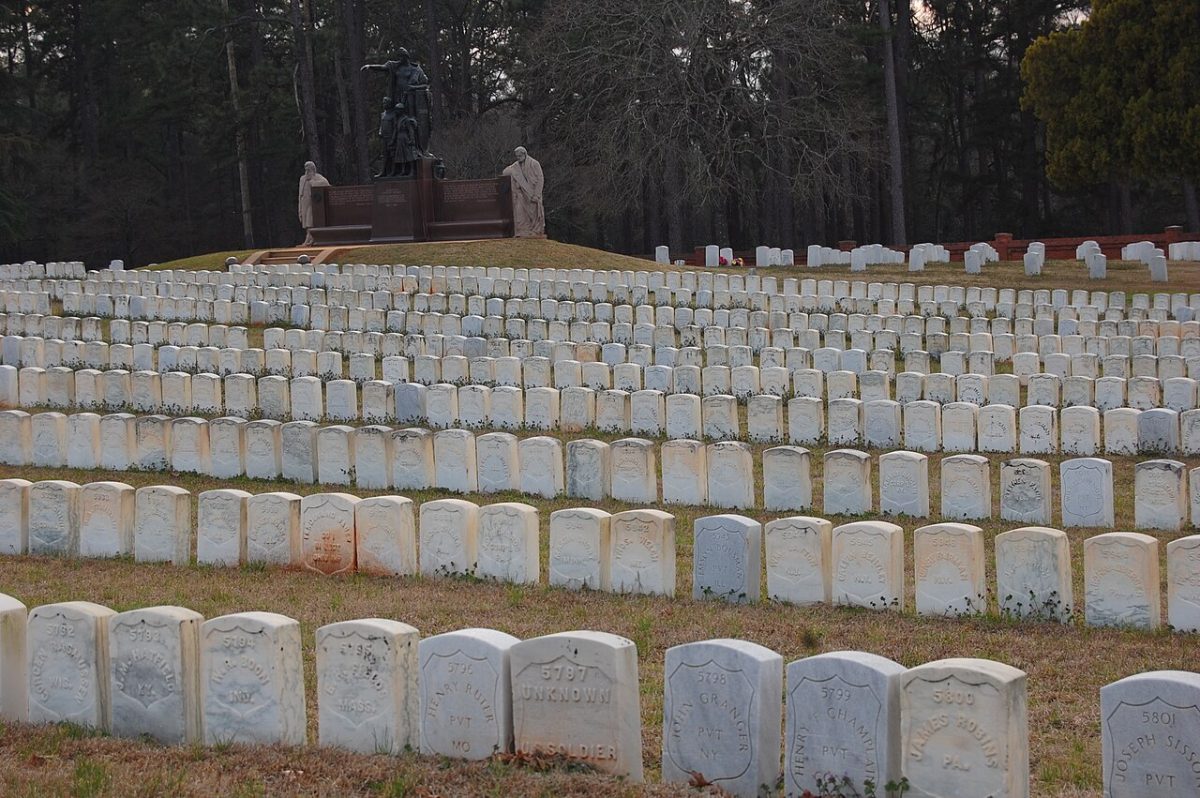 Rows of closely spaced white headstones climbing a slope at Andersonville National Cemetery, with a monument and pine woods behind