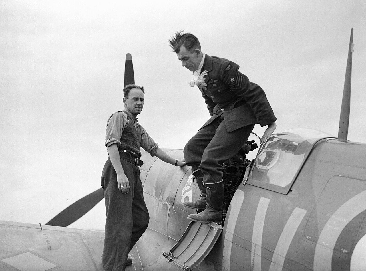 A pilot climbing out of a Spitfire cockpit onto the wing, helped by a man standing on the wing root