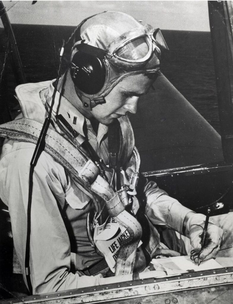 A young George H.W. Bush in flight helmet, goggles and life vest, writing on a clipboard in the cockpit of a TBM Avenger