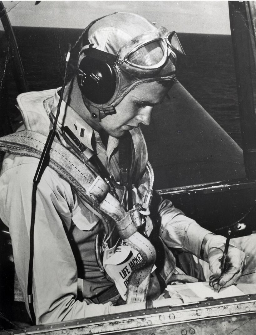 A young George H.W. Bush in flight helmet, goggles and life vest, writing on a clipboard in the cockpit of a TBM Avenger