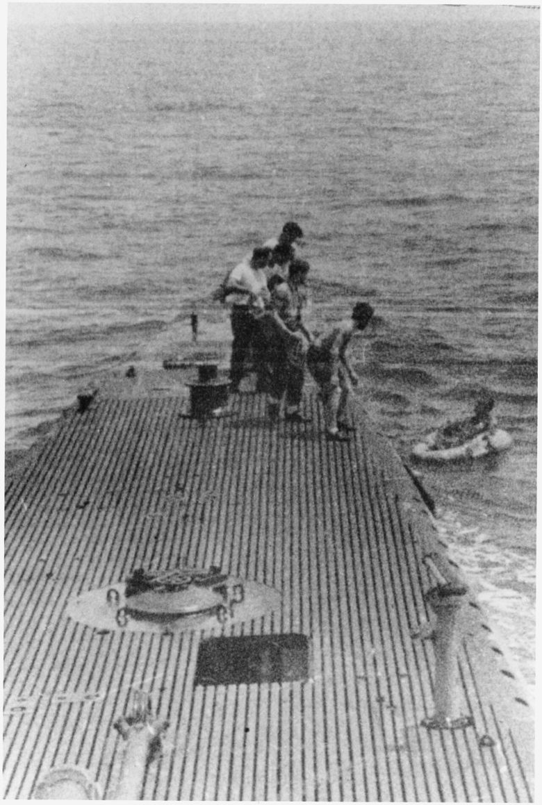 Crewmen on the deck of the submarine USS Finback reach toward a man in a small life raft alongside the hull