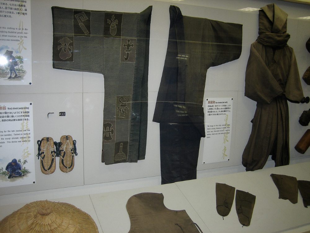 A museum display of historical ninja clothing: a reversible patterned jacket, dark trousers, straw sandals and a hooded brown outfit, with Japanese and English labels