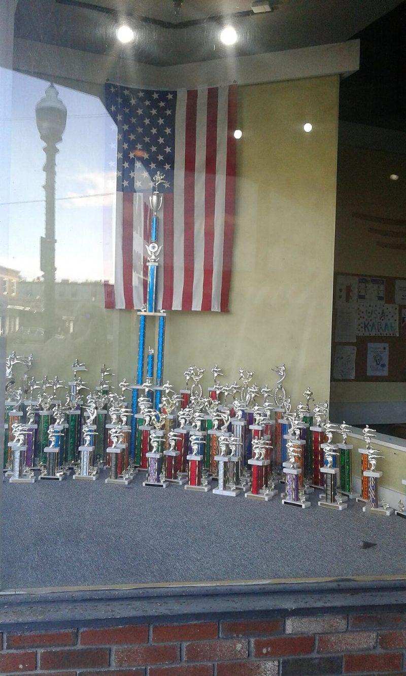 A shop window in Vermont filled with rows of nearly identical karate trophies, each topped with a small silver figure in a kicking pose