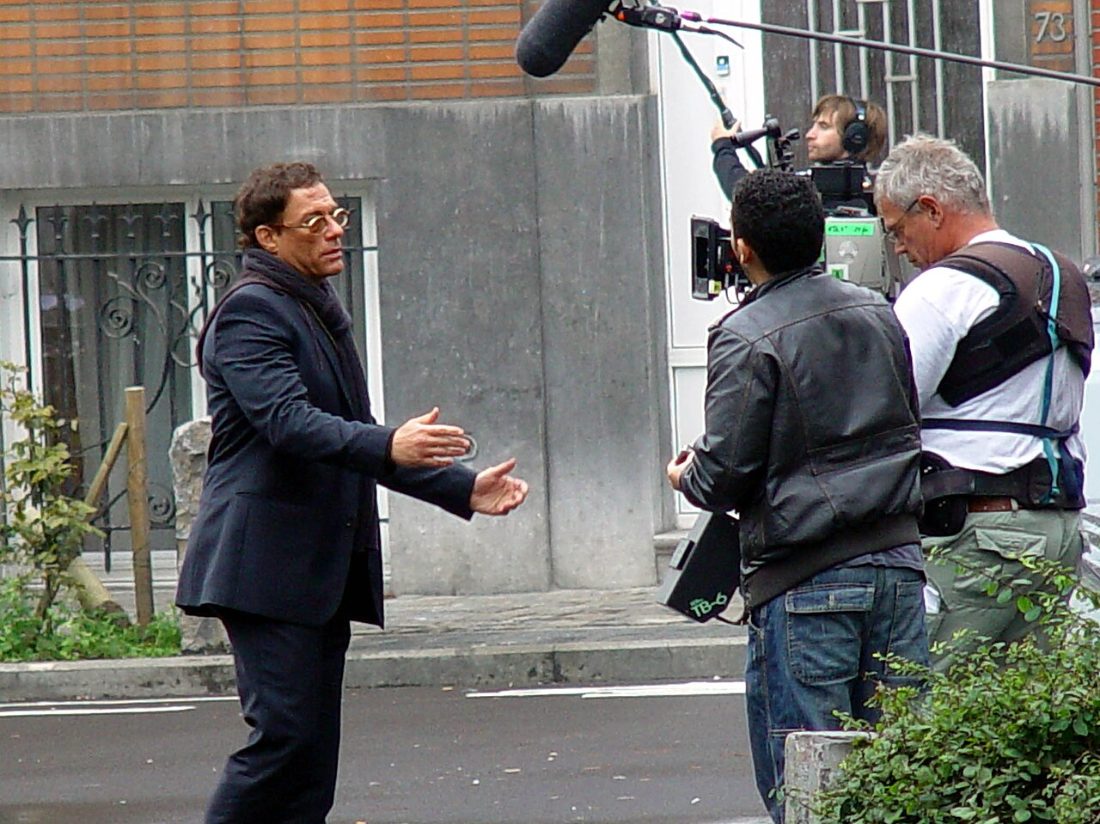 Jean-Claude Van Damme standing in a street in a dark suit and scarf, facing a camera operator and boom operator on a film set