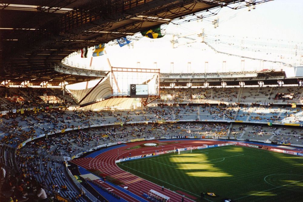 The inside of the Stadio delle Alpi in Turin in 1990, with a running track around the pitch, partly filled stands and Brazilian flags hanging from the roof