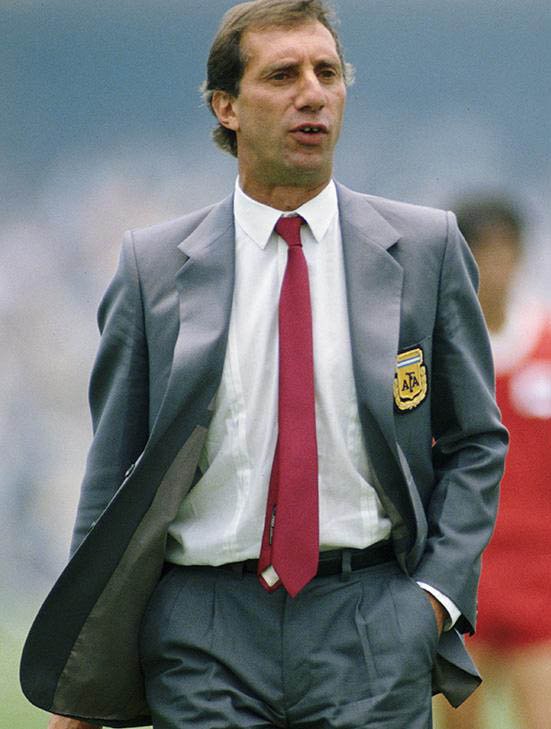 Carlos Bilardo in a grey suit with the Argentine federation badge and a red tie, walking on the touchline