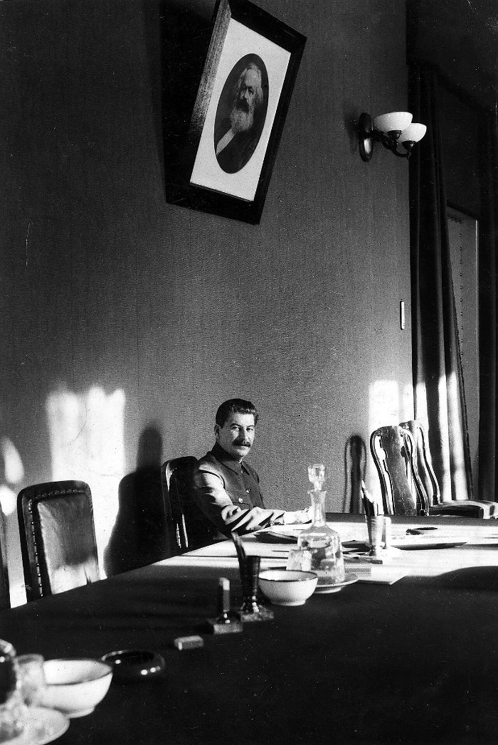 Joseph Stalin seated alone at the end of a long conference table in the Kremlin beneath a framed portrait of Karl Marx