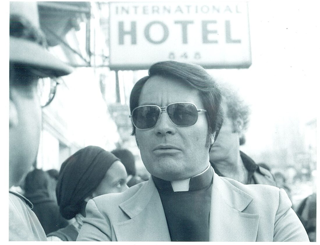Jim Jones in sunglasses and a clerical collar standing in a crowd beneath the International Hotel sign in San Francisco