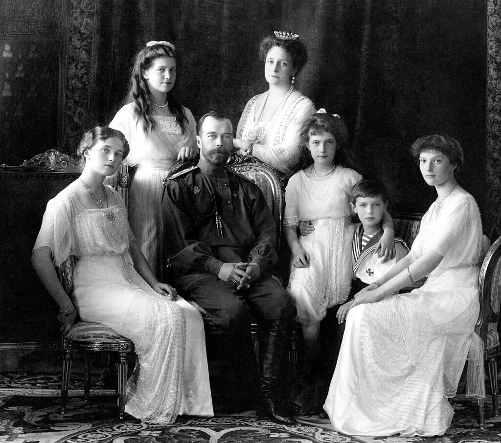 Royal family portrait: a man in a dark military uniform seated among six young women in white gowns in an ornate room.
