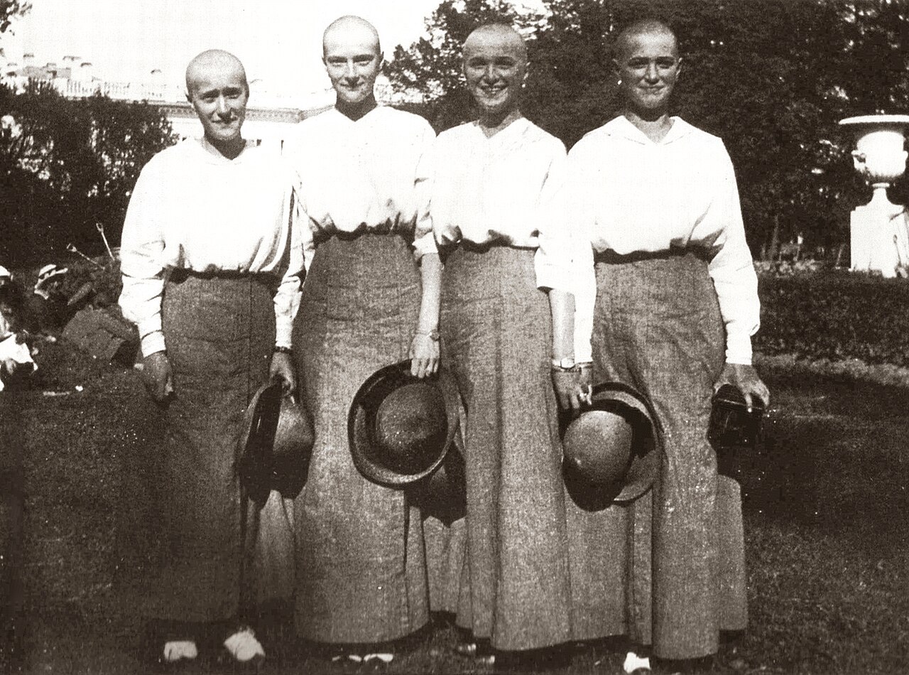 Four young women with shaved heads standing together in a garden, holding hats