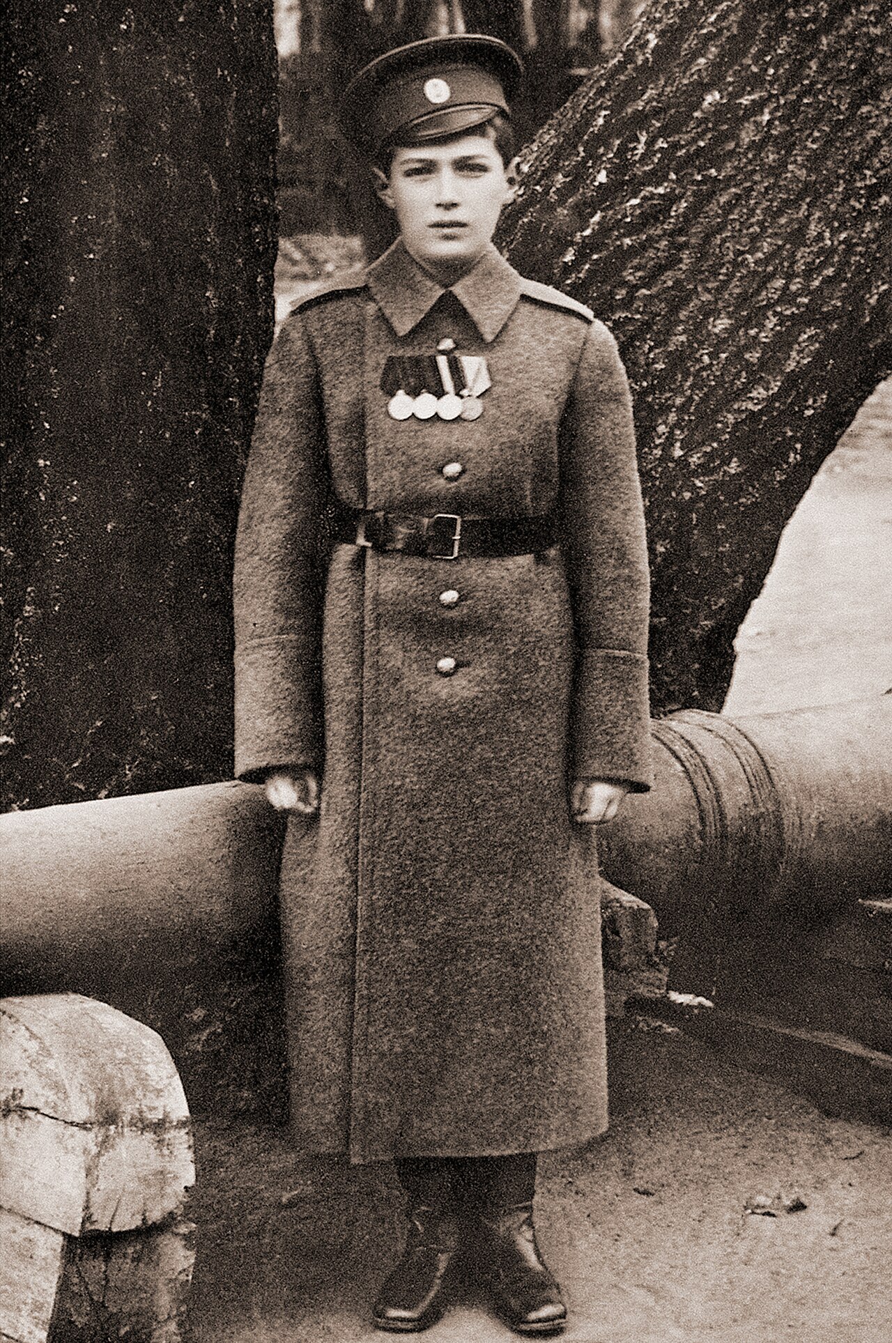 A boy in military uniform and peaked cap standing in front of a large gun barrel