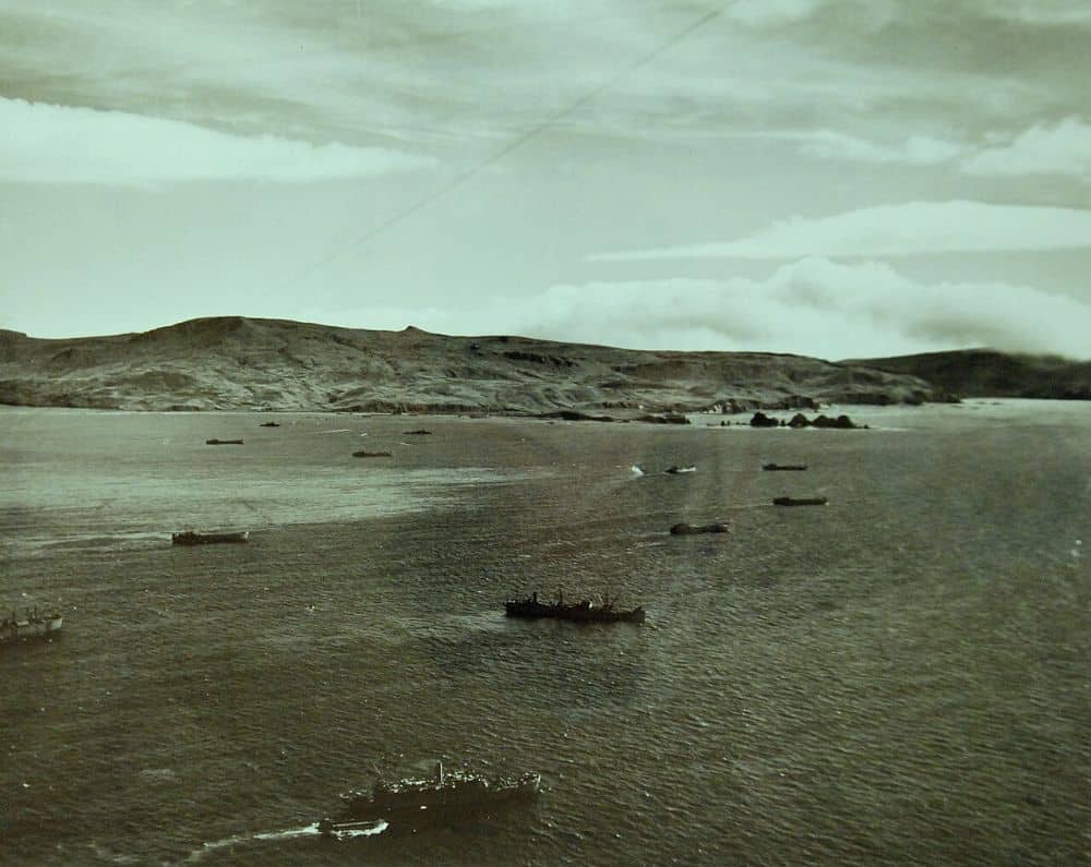Landing craft and U.S. Navy transports scattered across grey water off the bare, treeless ridgeline of Kiska Island, August 1943