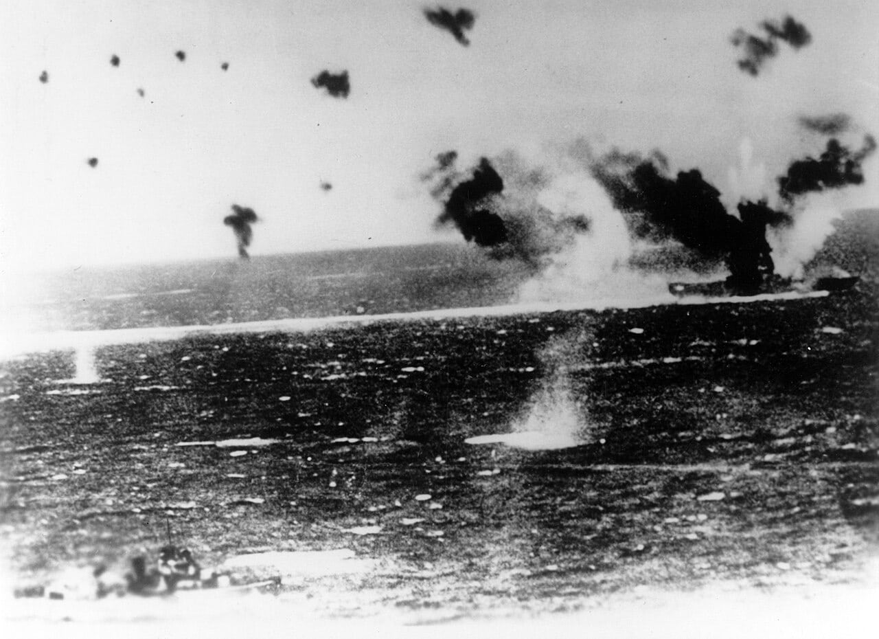 An American aircraft carrier under air attack, shell splashes and smoke around her