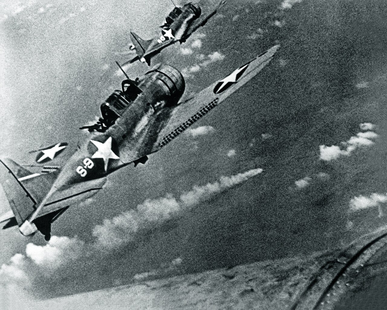 Two dive bombers flying above a burning Japanese heavy cruiser seen from high above