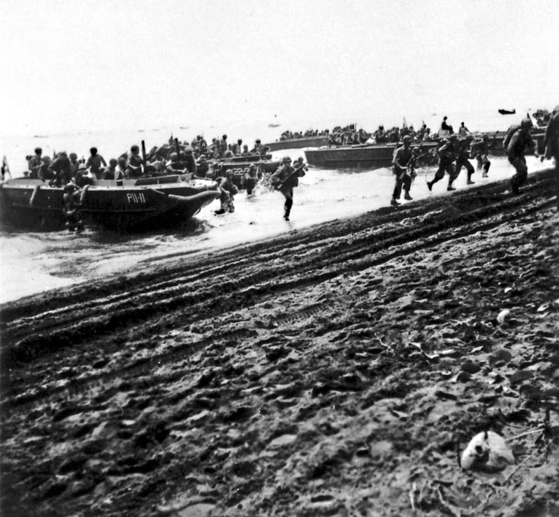 Marines wading through shallow water toward a palm-lined beach carrying packs and rifles
