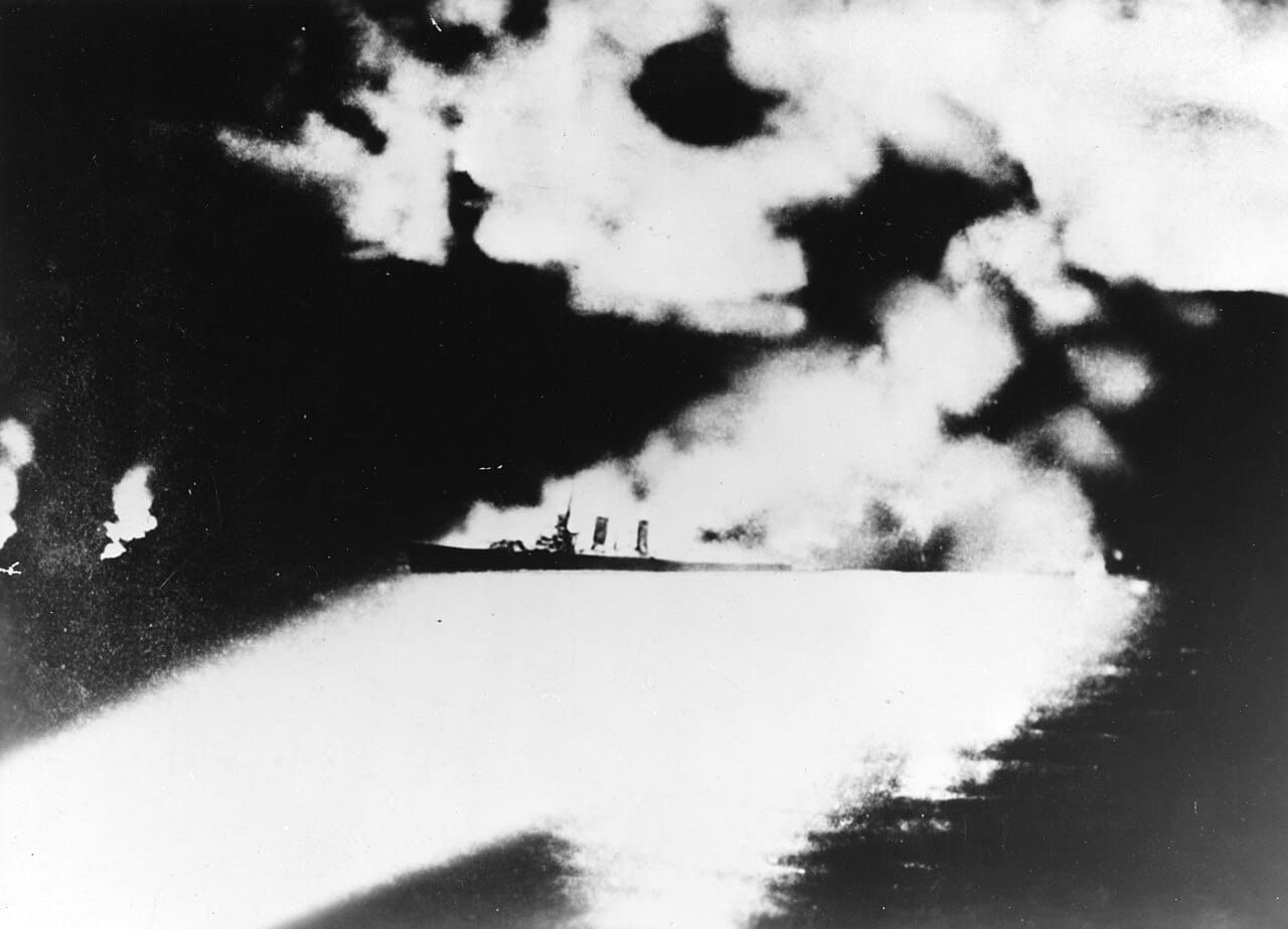 An American heavy cruiser lit by searchlights at night, under fire