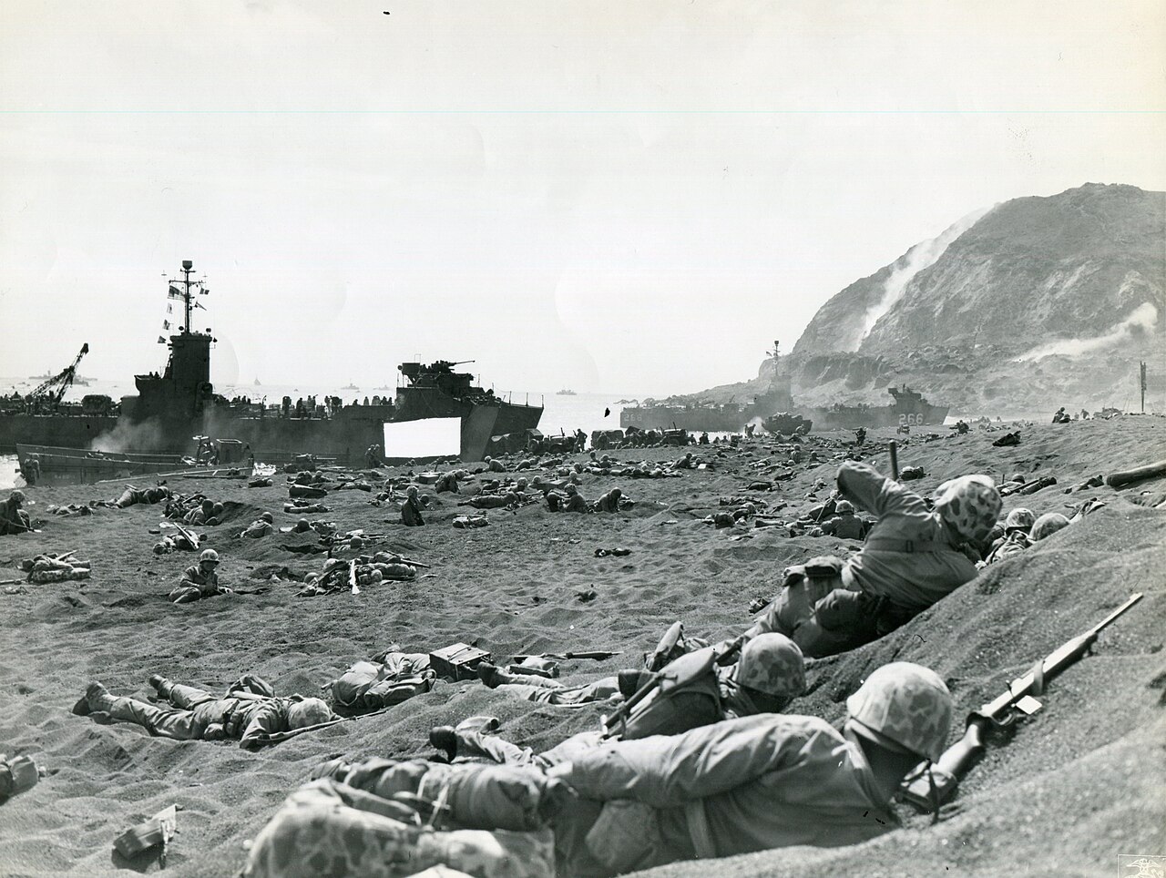 Marines burrowed into black volcanic sand while supplies are unloaded behind them