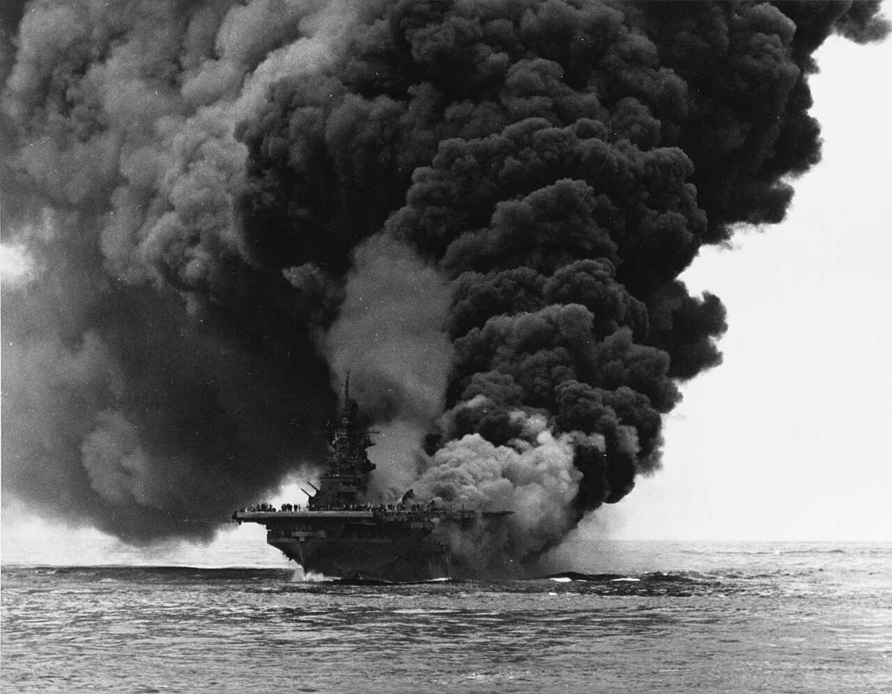 An aircraft carrier burning heavily with thick black smoke rolling off her deck
