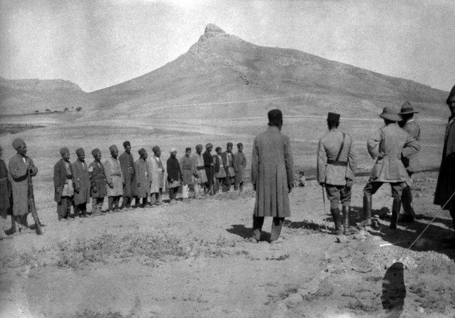 A line of Persian men in long coats standing in open country while several uniformed British officers face them, a rocky peak behind