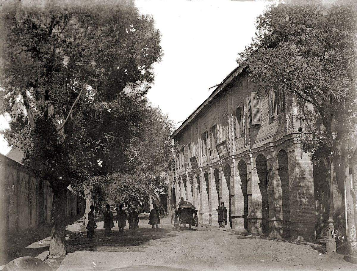 A tree-lined Tehran street around 1910, with arcaded brick buildings, a horse-drawn carriage and a handful of pedestrians