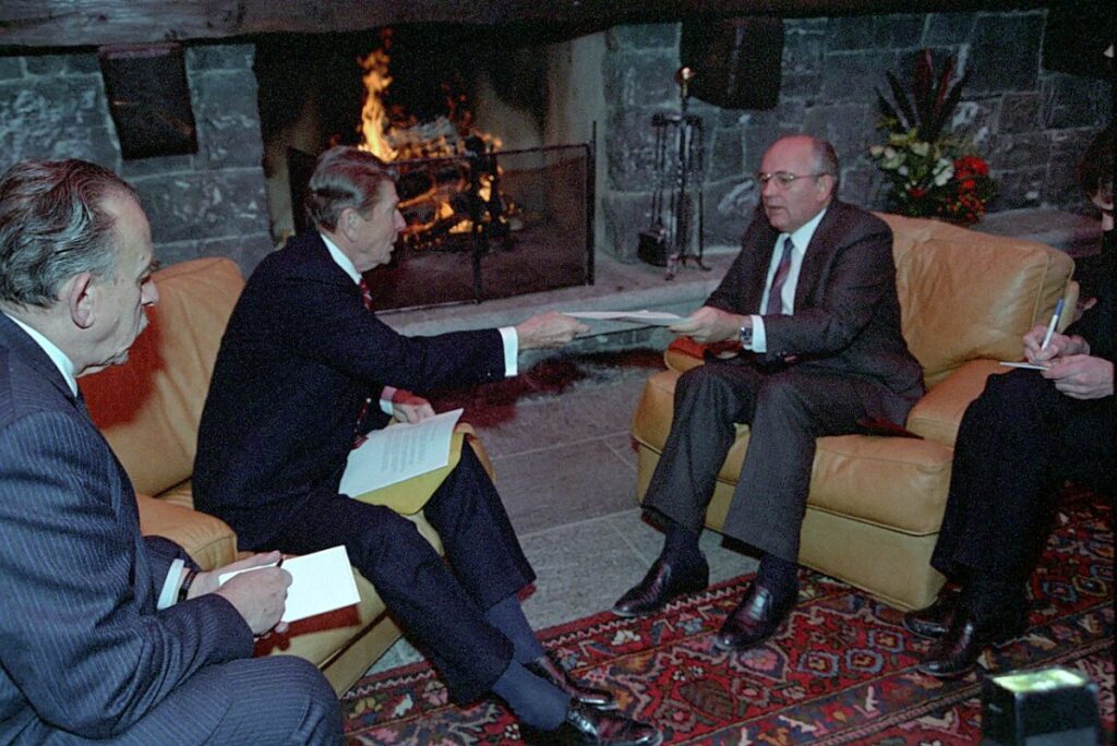 Reagan and Gorbachev seated in armchairs in front of a stone fireplace at the Fleur d'Eau pool house in 1985, each with an interpreter taking notes