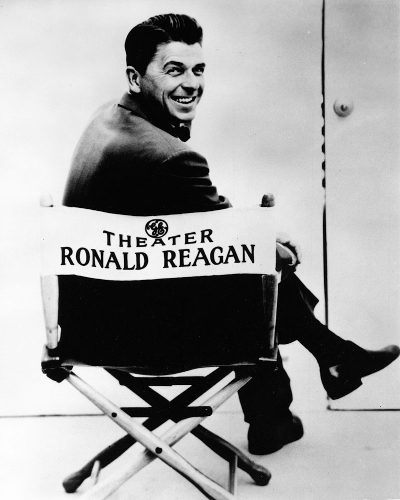 A 1950s publicity photograph of Ronald Reagan grinning over his shoulder while sitting in a director's chair printed GE THEATER RONALD REAGAN