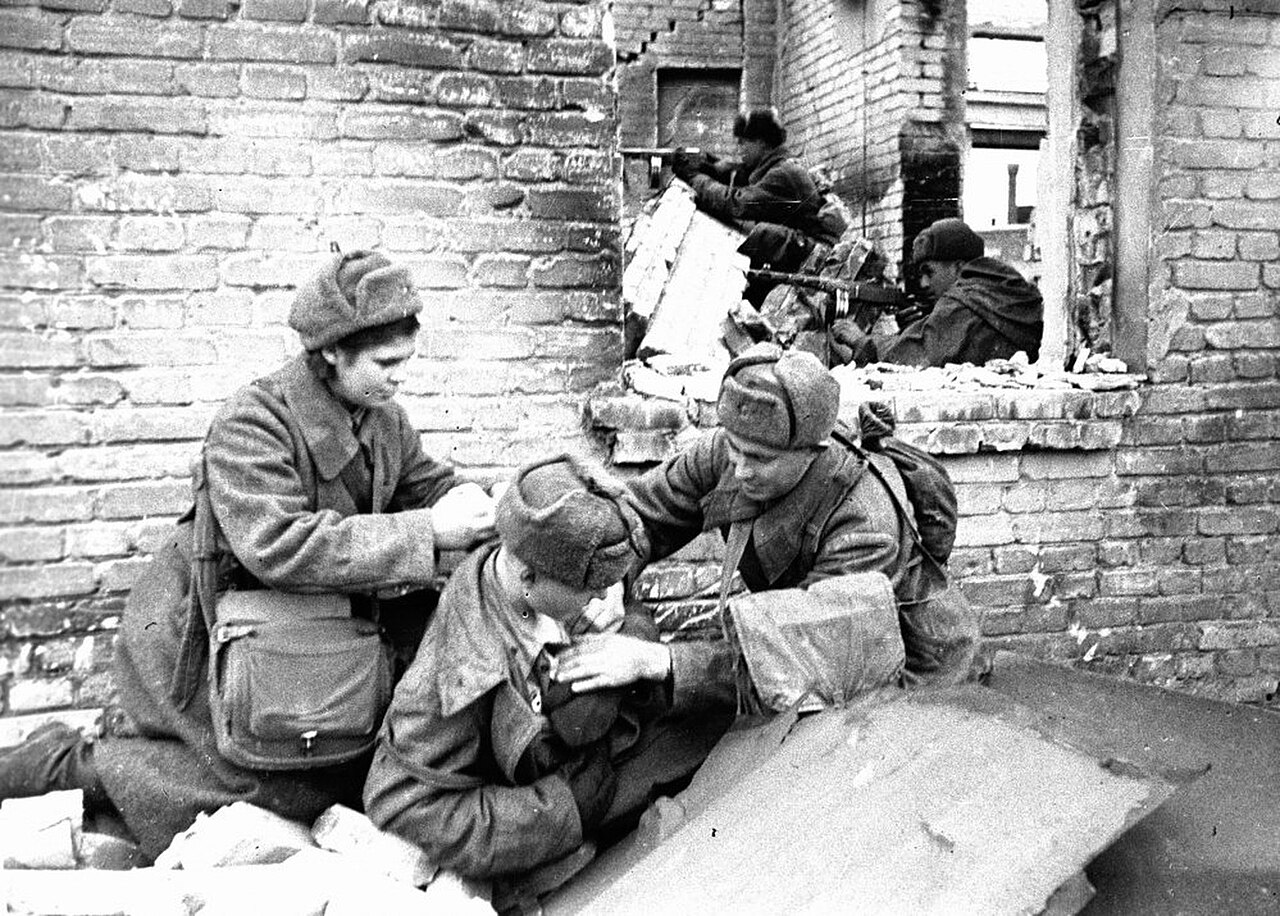 Two Soviet medical orderlies bandaging a wounded soldier beside a brick wall