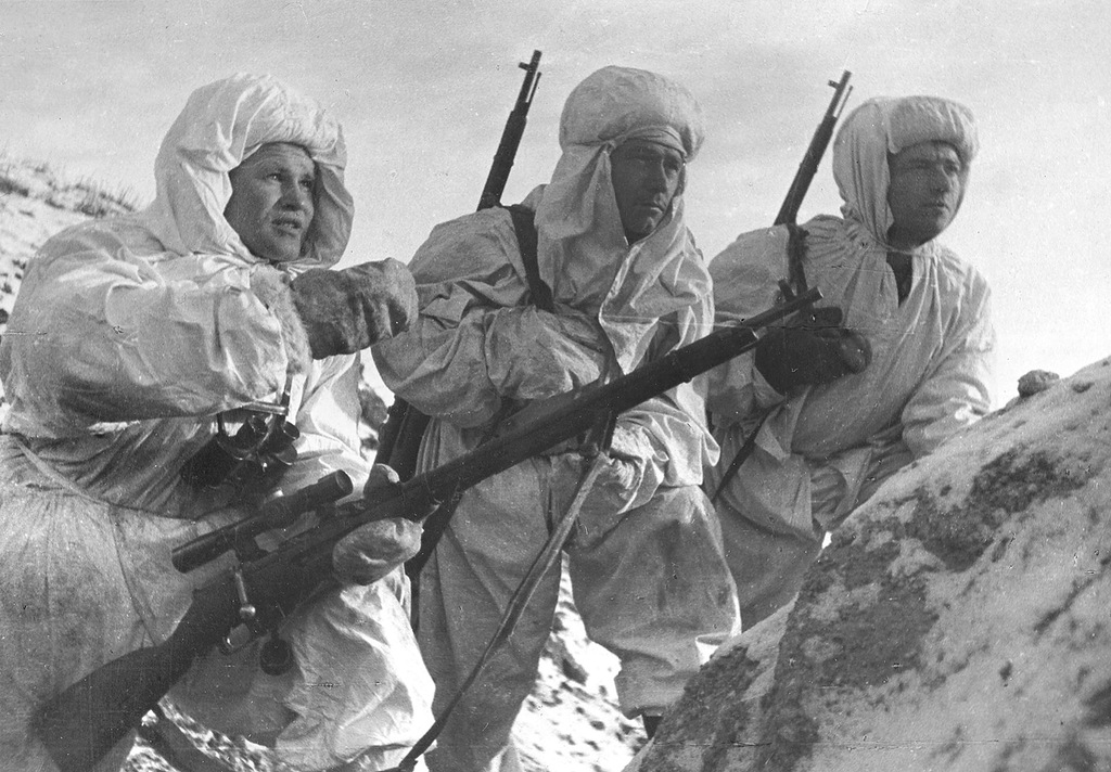 A group of snipers in white winter camouflage with scoped rifles, one man briefing the others