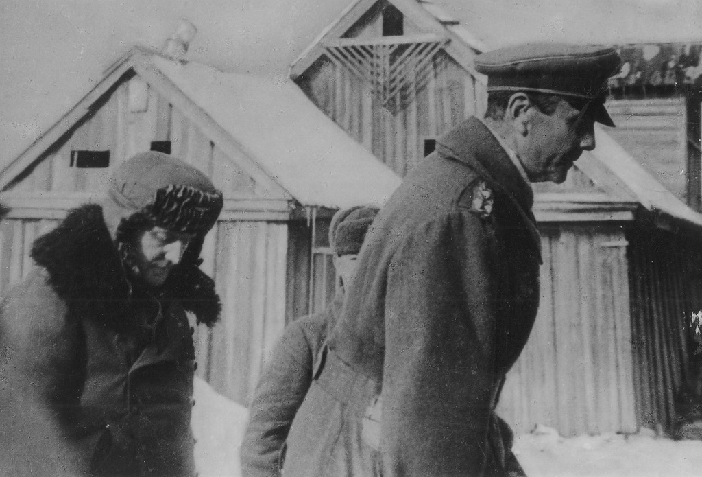 A German officer in a greatcoat walking through snow past wooden huts under escort