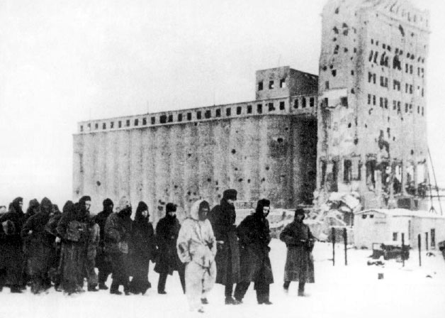 A long column of prisoners walking through snow past a huge damaged grain elevator