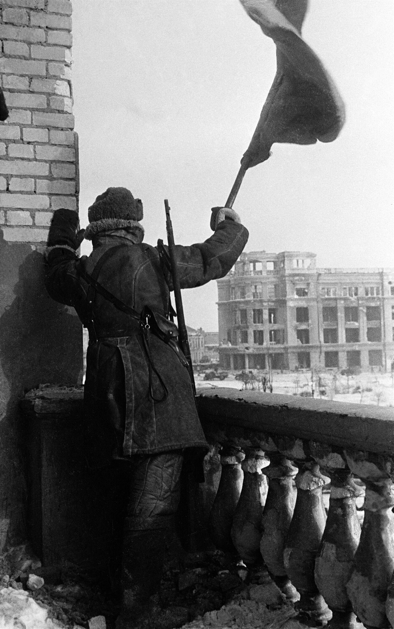 A Red Army soldier on a shattered balcony raising a flag above a ruined square