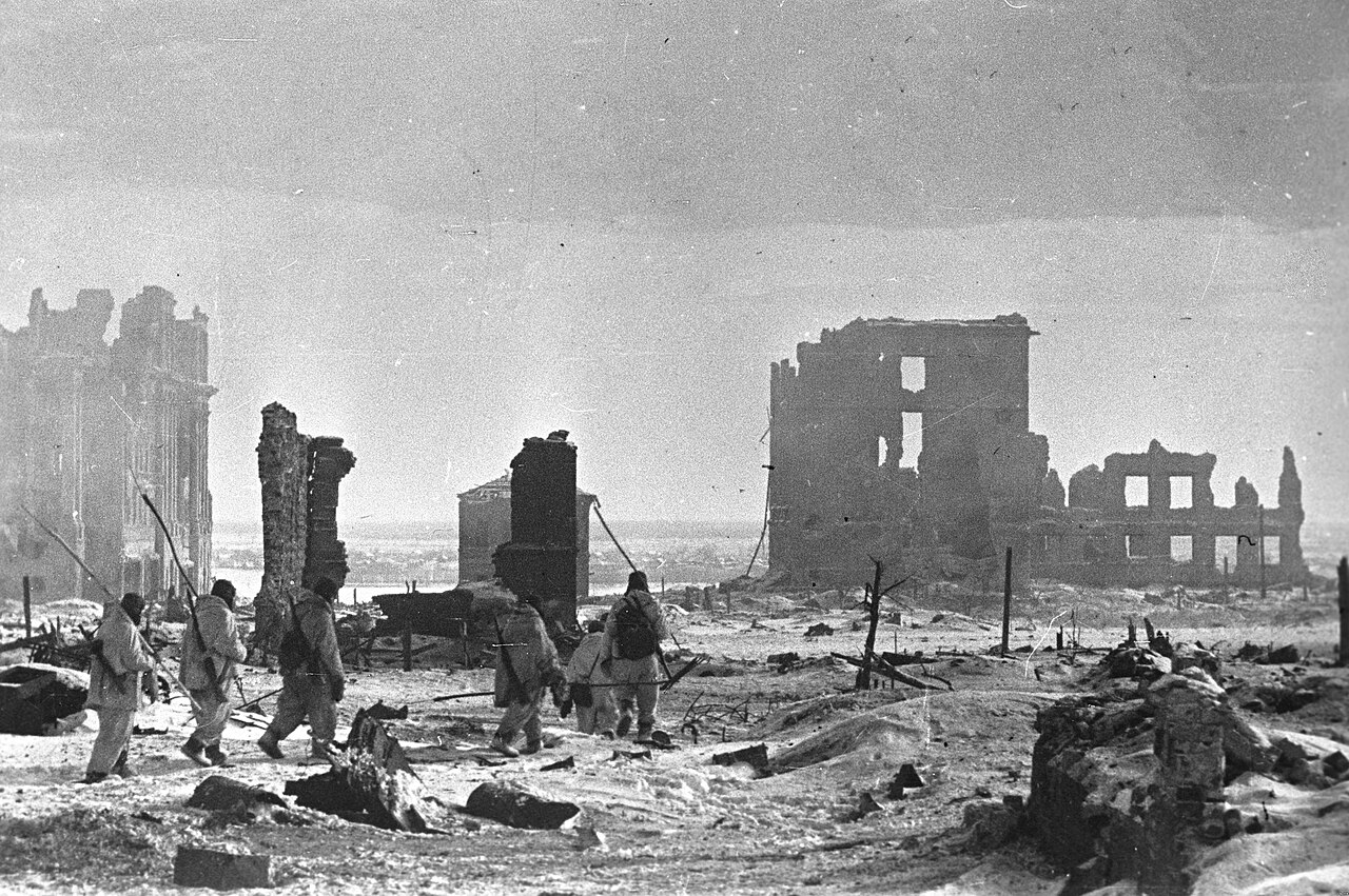 Figures crossing open, snow-covered ground between burnt-out buildings in a city centre