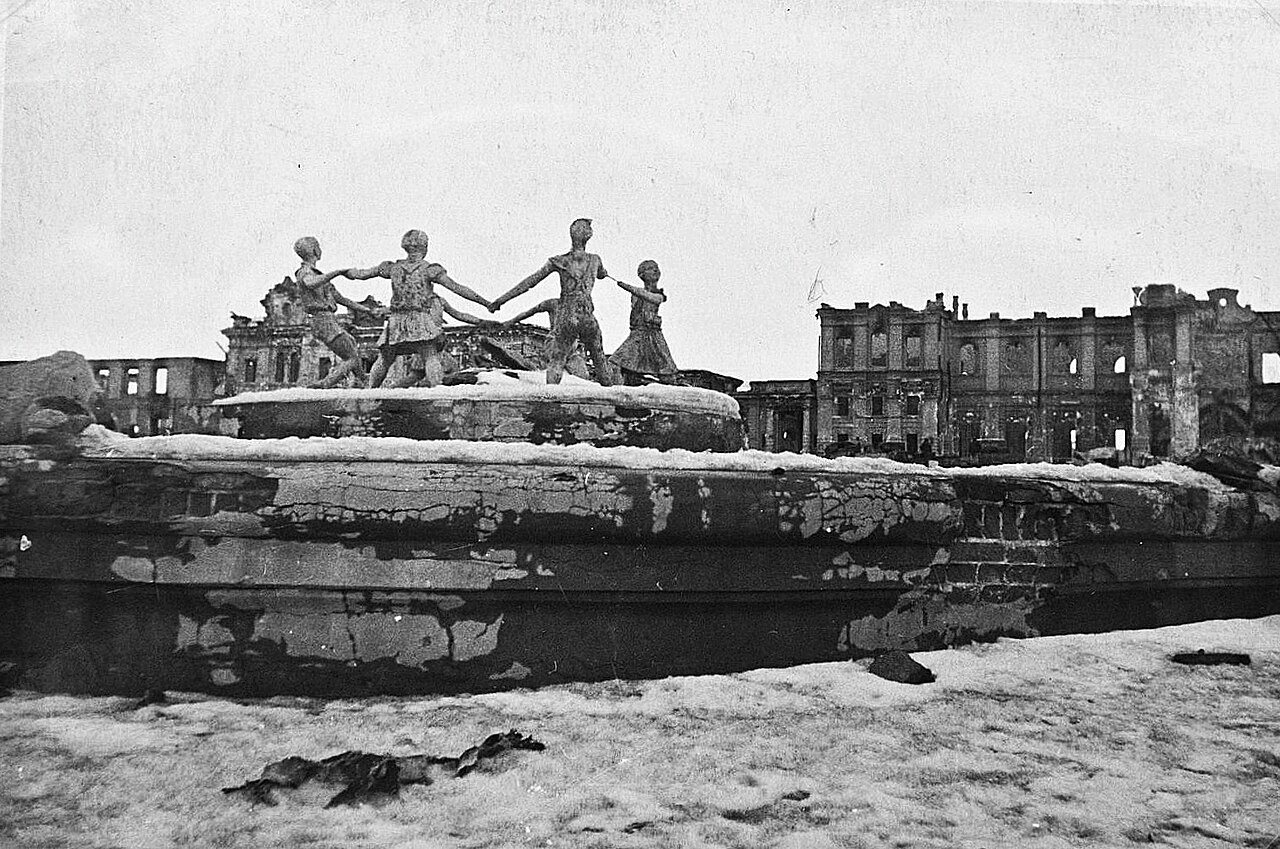 A fountain sculpture of children dancing in a ring, standing intact in front of ruined buildings