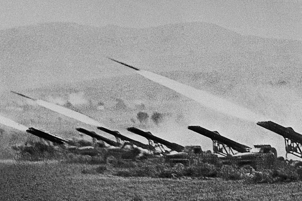 A battery of rocket launchers firing, smoke trails streaking across open ground