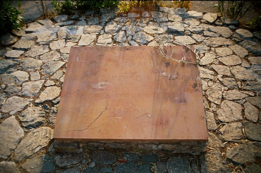 A flat stone slab set into a cobbled surface at Thermopylae, carved with the Greek text of Simonides' epitaph for the Spartan dead