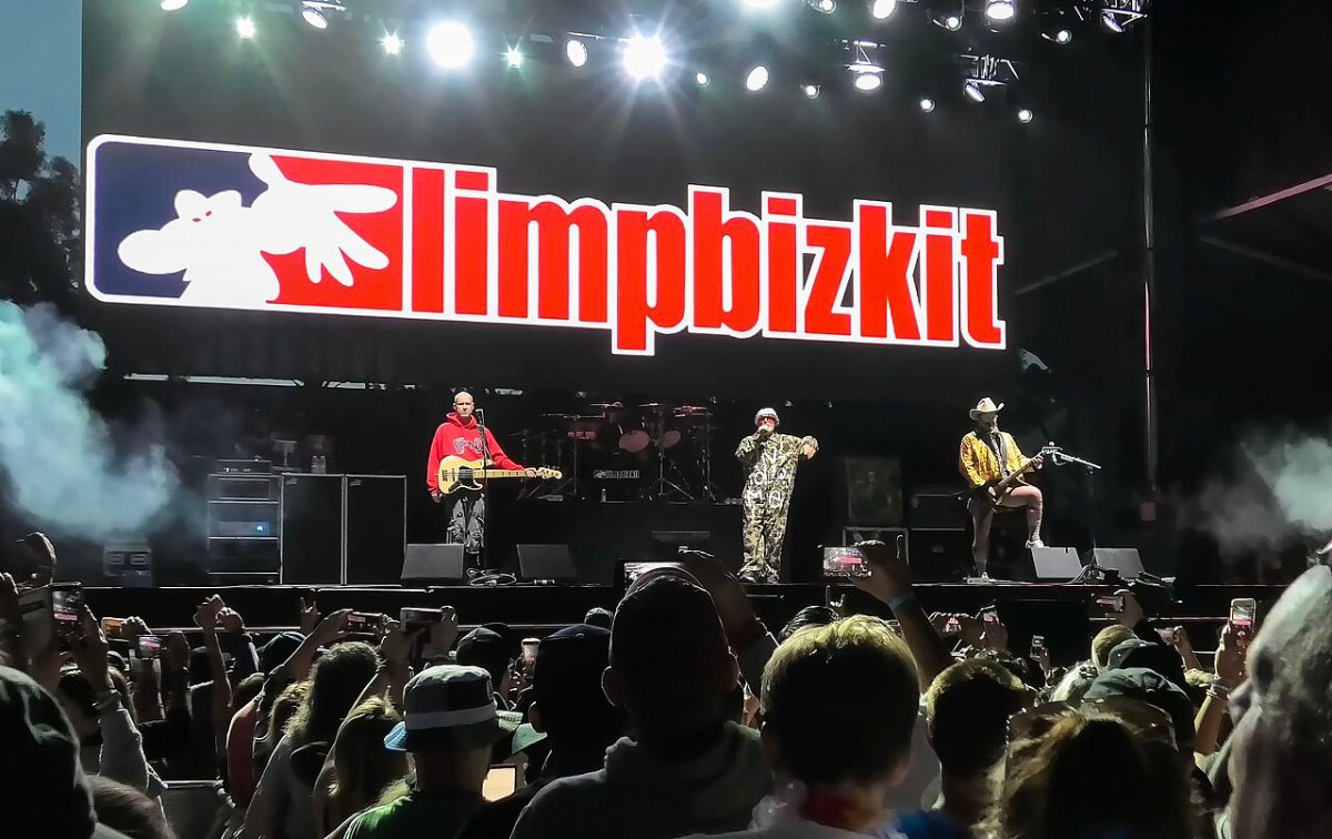 Limp Bizkit performing on a floodlit festival stage beneath a huge illuminated band logo, with a crowd of raised phones in the foreground