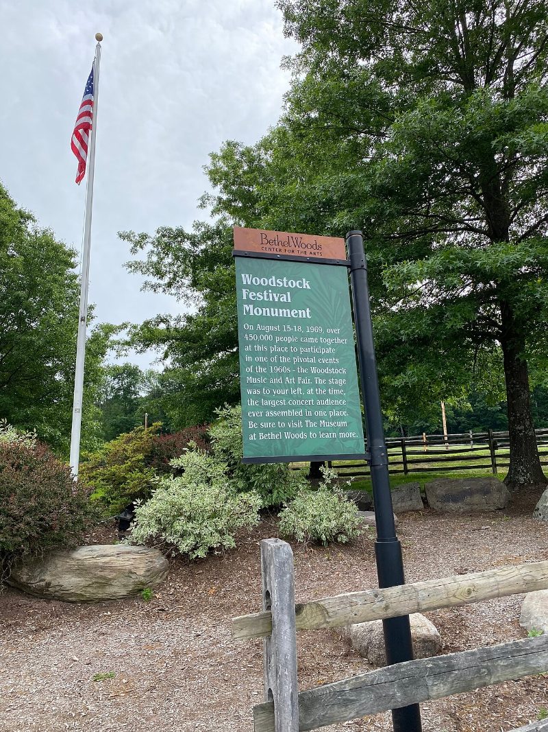 A green interpretive sign reading Woodstock Festival Monument beside a flagpole and split-rail fence at Bethel Woods, New York